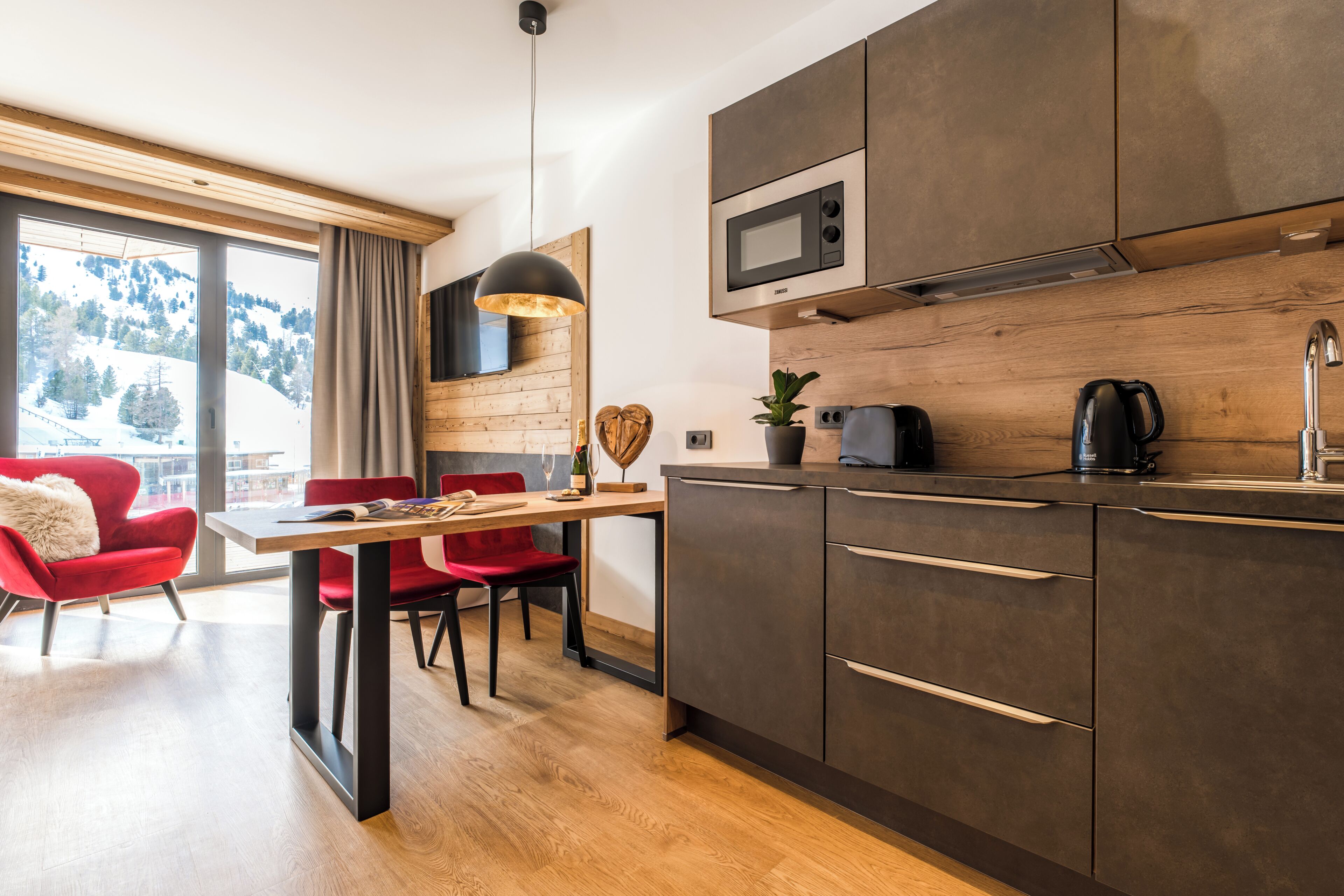 Una cocina moderna con armarios oscuros, microondas y cafetera. Hay una zona de comedor con mesa y sillas, y un ventanal con vistas a un paisaje nevado.
