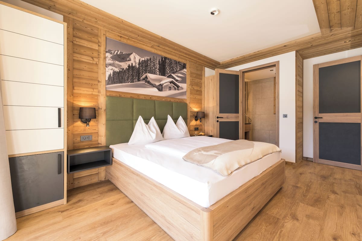 A bedroom with a wooden headboard, a bed with white bedding, a nightstand with a lamp, and a painting of a snowy mountain scene.