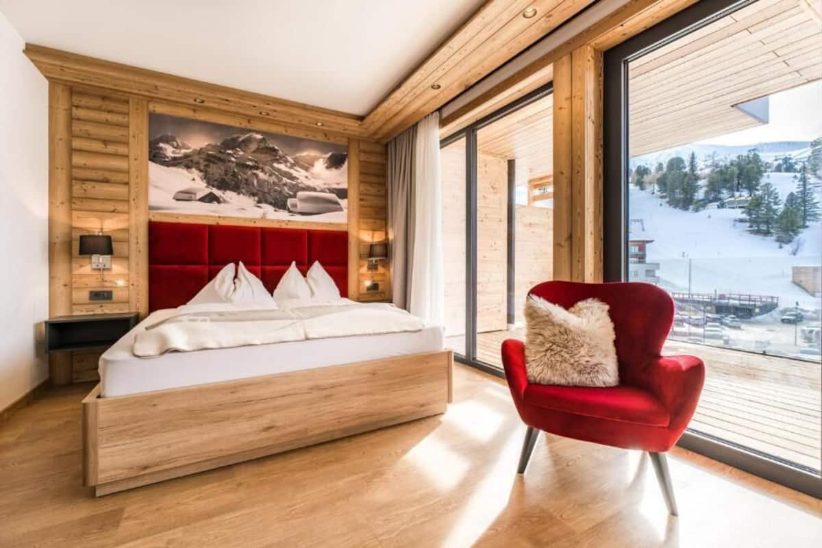 A modern bedroom with a wooden headboard, a large window with a mountain view, a red armchair, and a bed with white bedding.