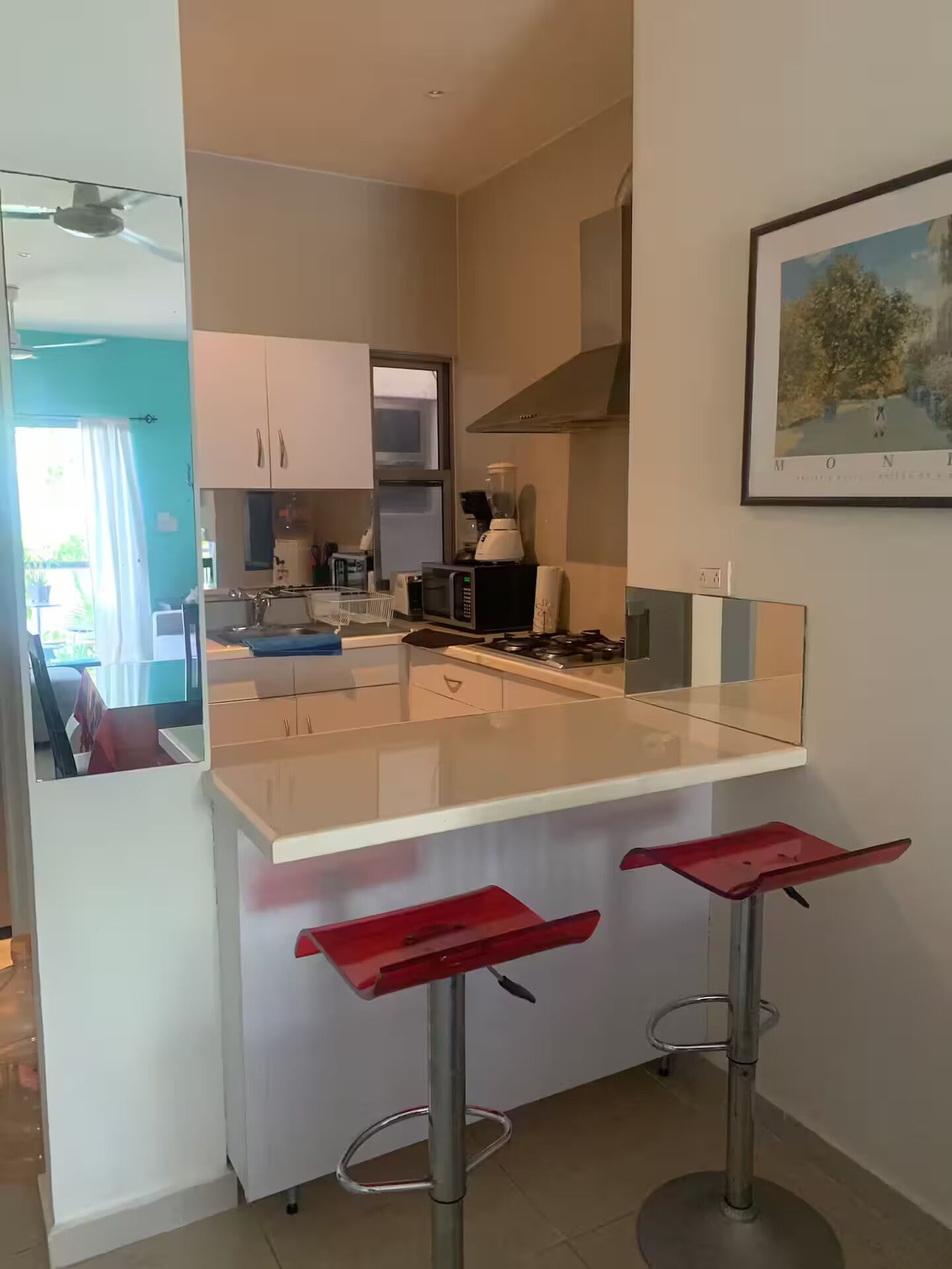 A modern kitchen with a central island, a microwave, a blender, and a gas stove. There are two red bar stools and a framed picture on the wall.