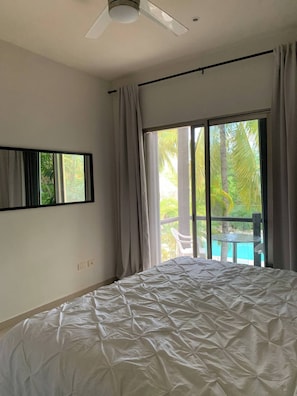 A bedroom with a large bed, a ceiling fan, a window with curtains, and a view of a pool and palm trees. - Beautiful condo by the beach (Playa del Carmen)