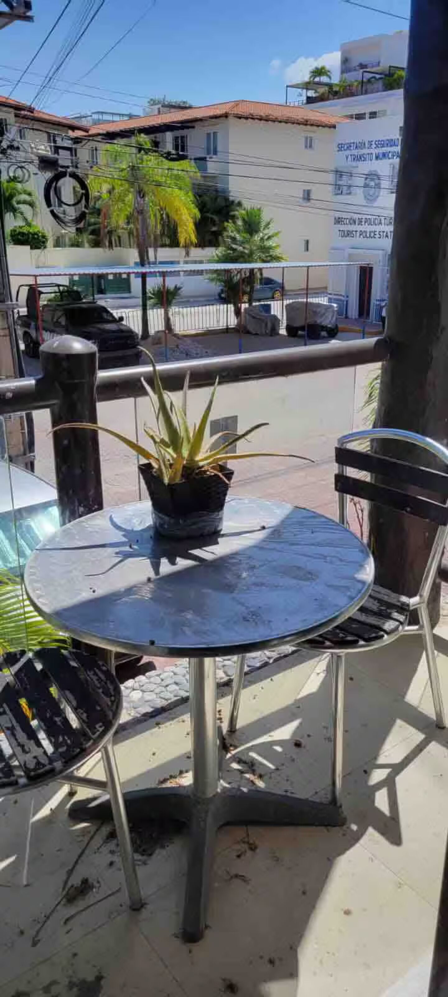A round metal table with a potted plant, two metal chairs, and a railing overlooking a street with buildings and parked cars.
