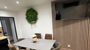 A modern dining area with a rectangular table, six chairs, a vase with flowers, a wall-mounted TV, and a vertical wooden panel. - Elegant T2 Design & Comfort - Premium Getaway (Calais)