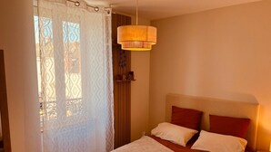 Ein Schlafzimmer mit einem Bett, einem Fenster mit gemustertem Vorhang, einer Hängelampe und einem Regal mit Dekoration.