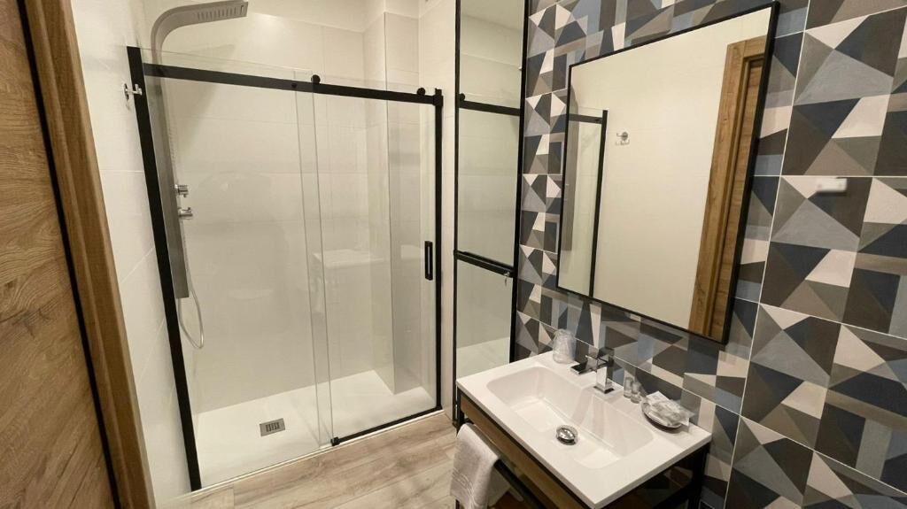 Um banheiro moderno com chuveiro em gabinete de vidro, pia branca e espelho com detalhes de azulejos geométricos.