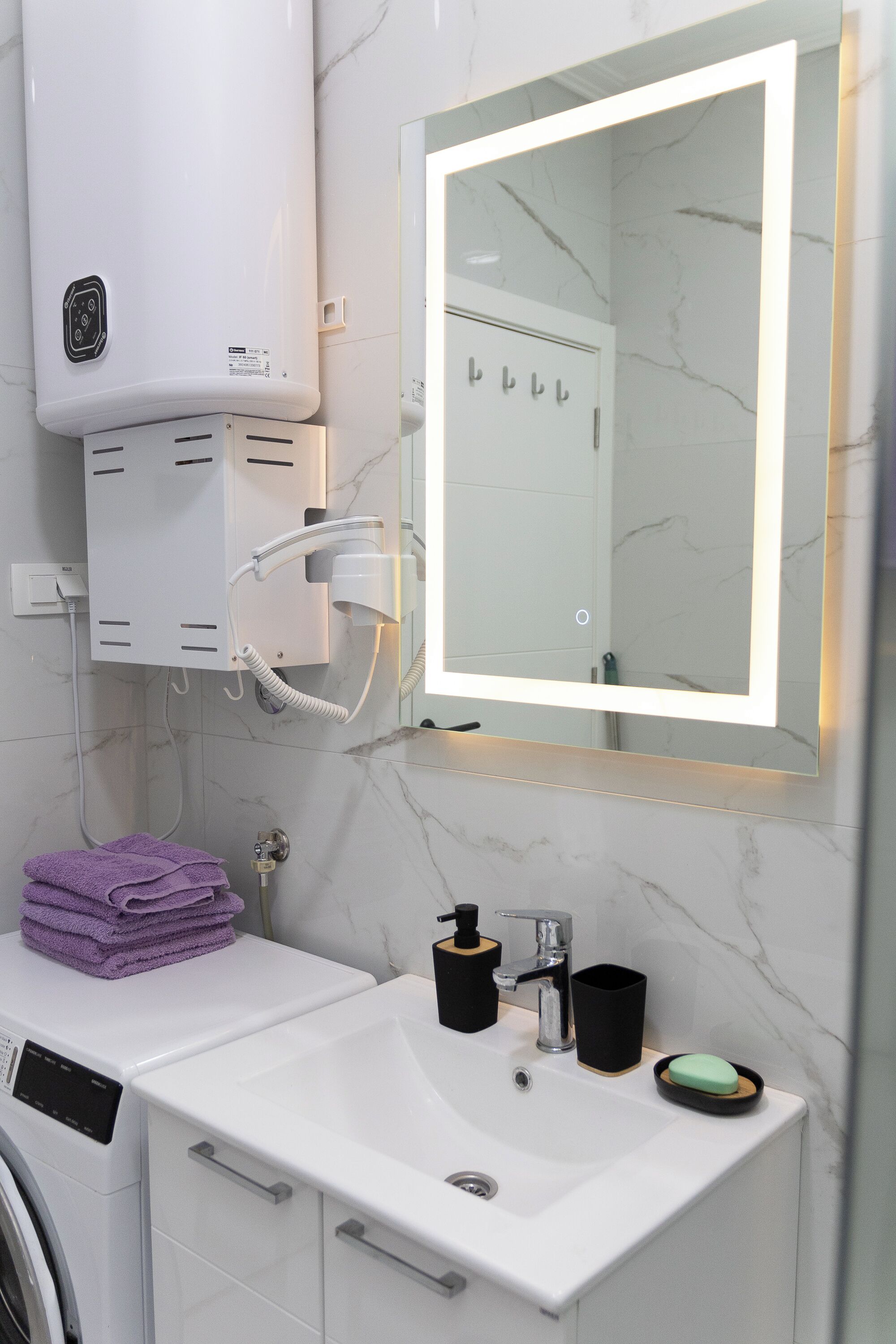 A modern bathroom with a built-in washing machine, a mirror with integrated lighting, a sink with a soap dish, and a towel stack.