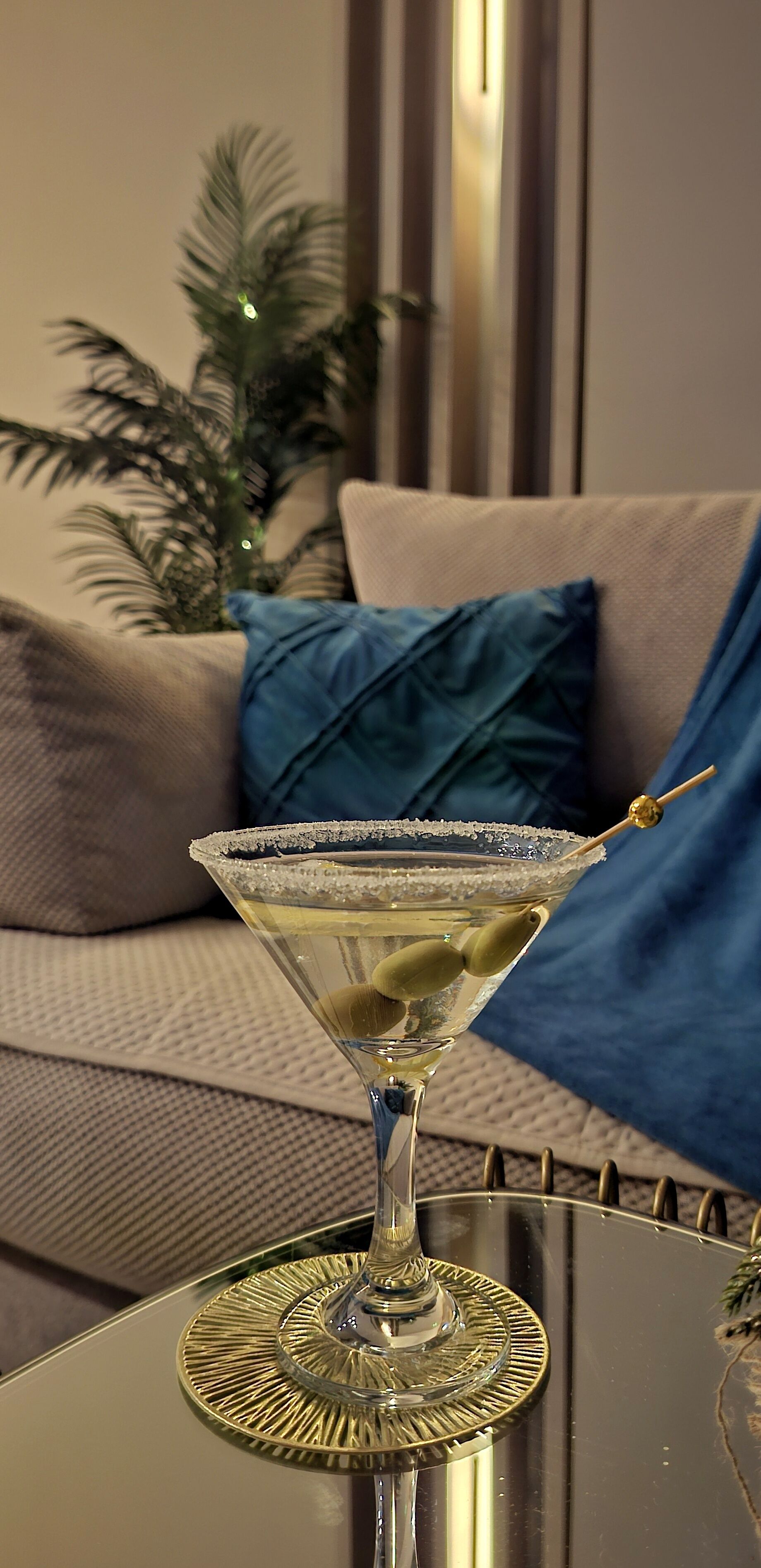 A martini glass on a table with a decorative coaster, a sofa with a blue pillow, and a potted plant in the background.