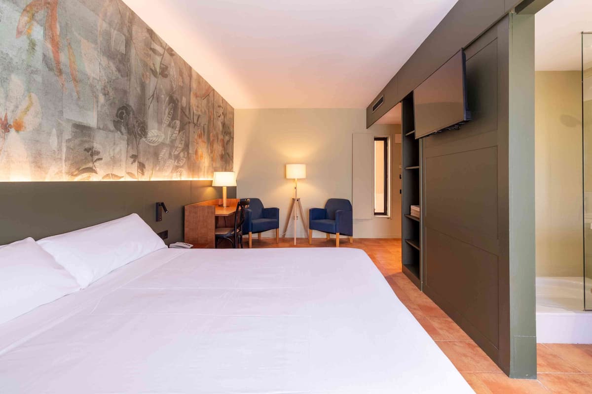 Une chambre d’hôtel avec un grand lit, un bureau avec deux chaises, un téléviseur fixé au mur et un grand tableau abstrait.