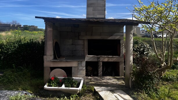 Une structure de four extérieur en pierre, dotée d’une cheminée, entourée de verdure et surplombant un ciel dégagé.