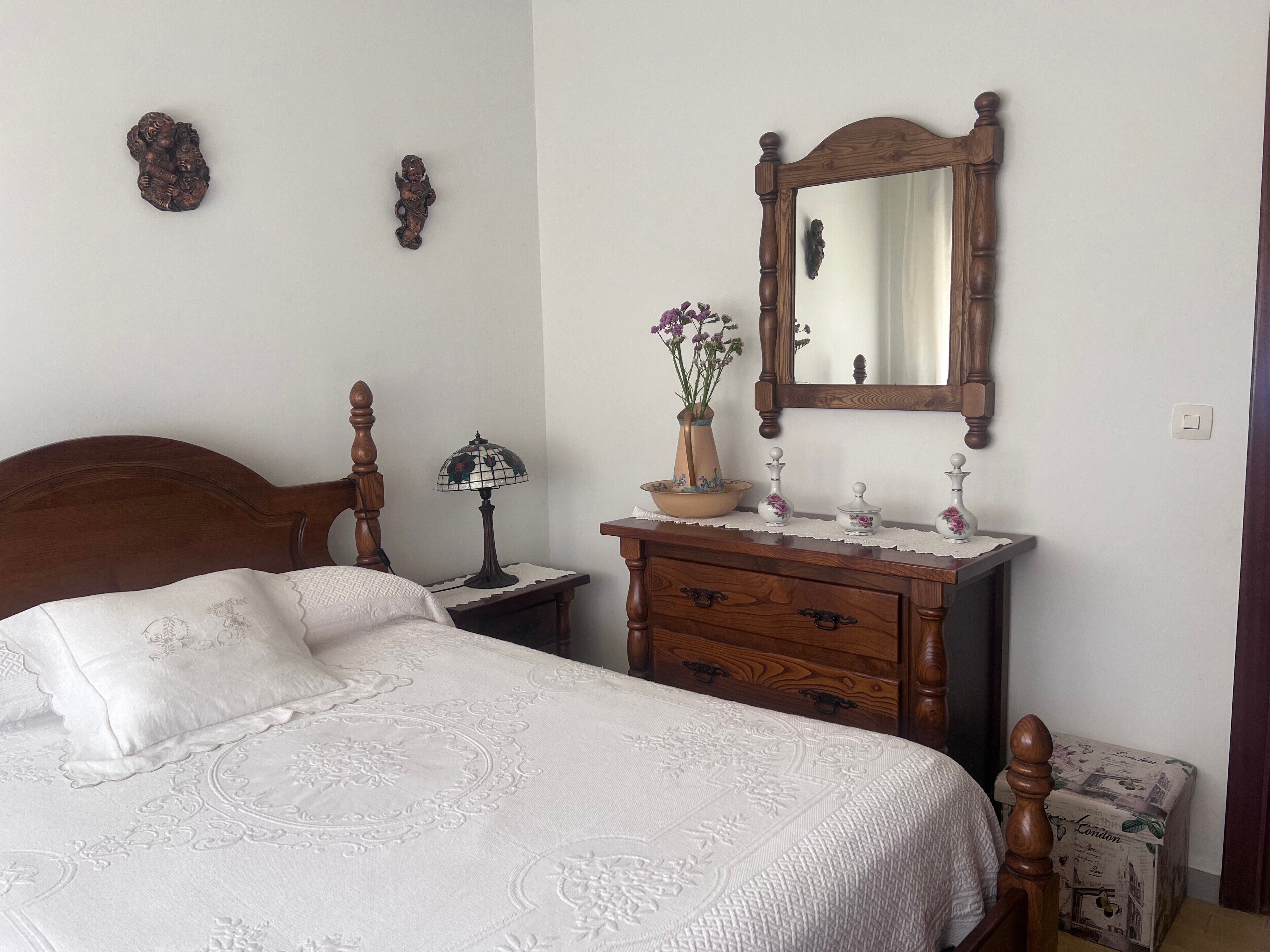 A bedroom with a wooden bed, a nightstand with a lamp, a dresser, a mirror, and a vase with flowers.