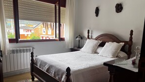 Une chambre à coucher avec un lit en bois, du linge de lit blanc, une fenêtre avec des stores, un radiateur et une table de chevet avec une lampe.