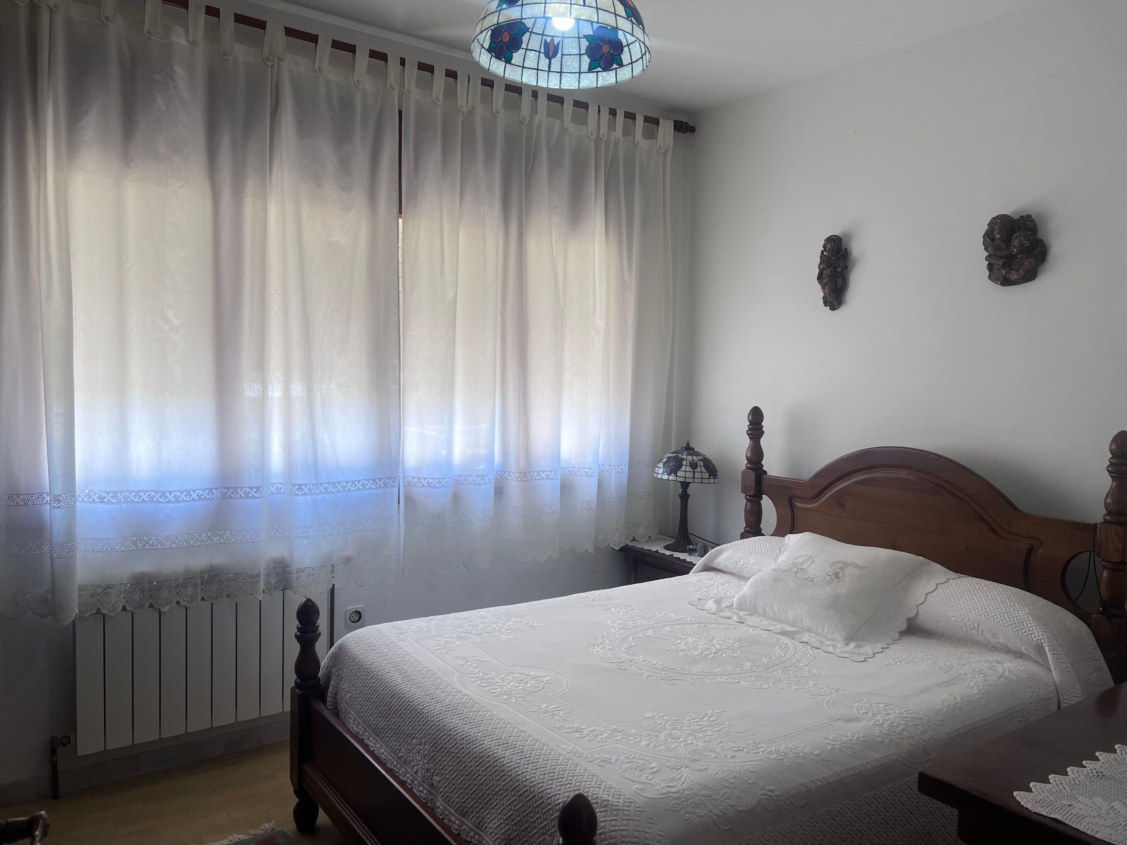 Un dormitorio con una cama de madera, ropa de cama blanca, una cortina de encaje, una lámpara de techo de vitral y un radiador.
