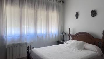 Un dormitorio con una cama de madera, ropa de cama blanca, una cortina de encaje, una lámpara de techo de vitral y un radiador.