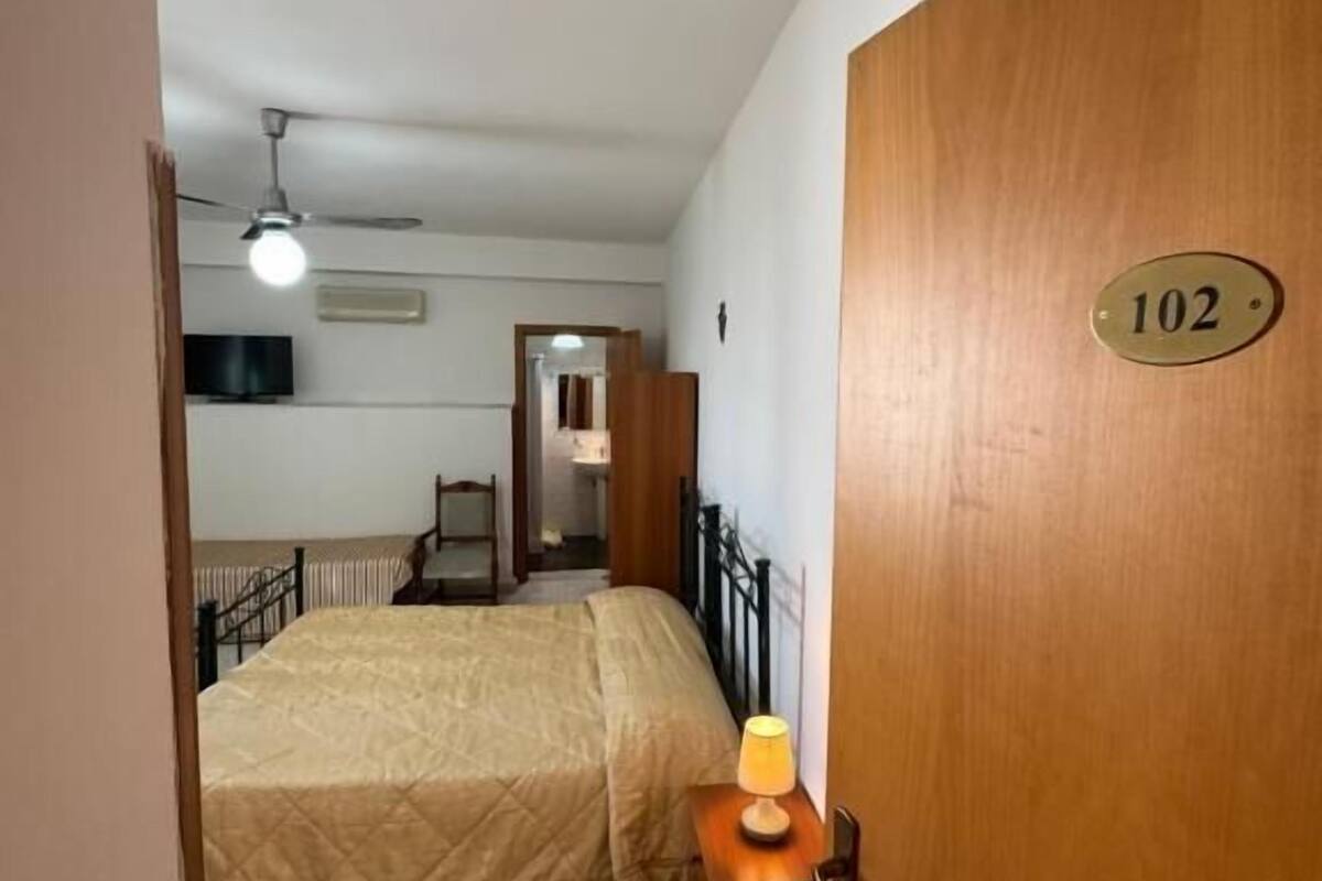Une chambre d’hôtel avec un lit, une table de chevet avec une lampe, un ventilateur de plafond et une porte sur laquelle est indiqué le numéro de la chambre.