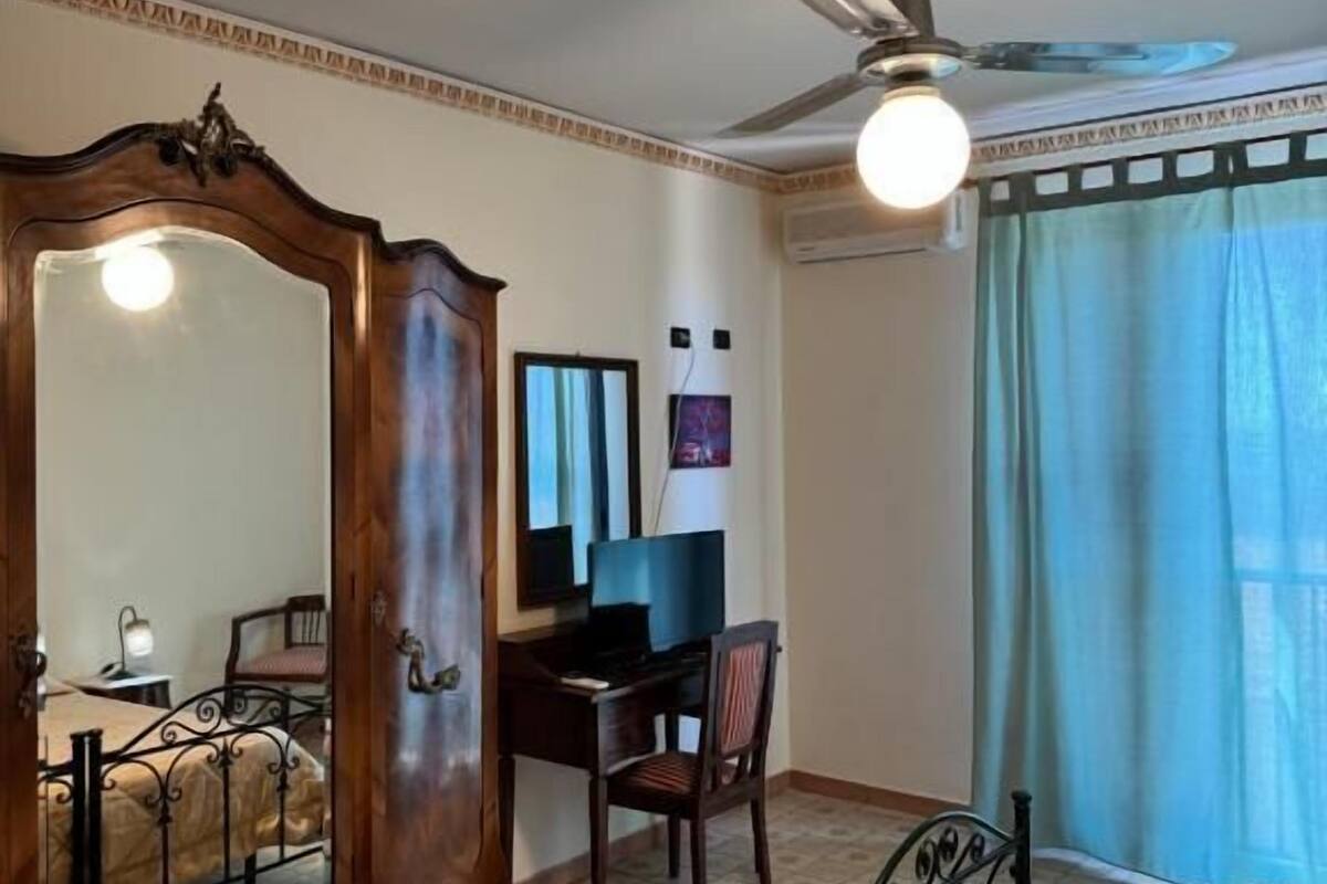Une chambre à coucher comprenant un lit, un bureau, une chaise, une armoire, un ventilateur de plafond et une fenêtre avec des rideaux.