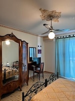 A bedroom with a bed, a desk, a chair, a wardrobe, a ceiling fan, and a window with curtains.