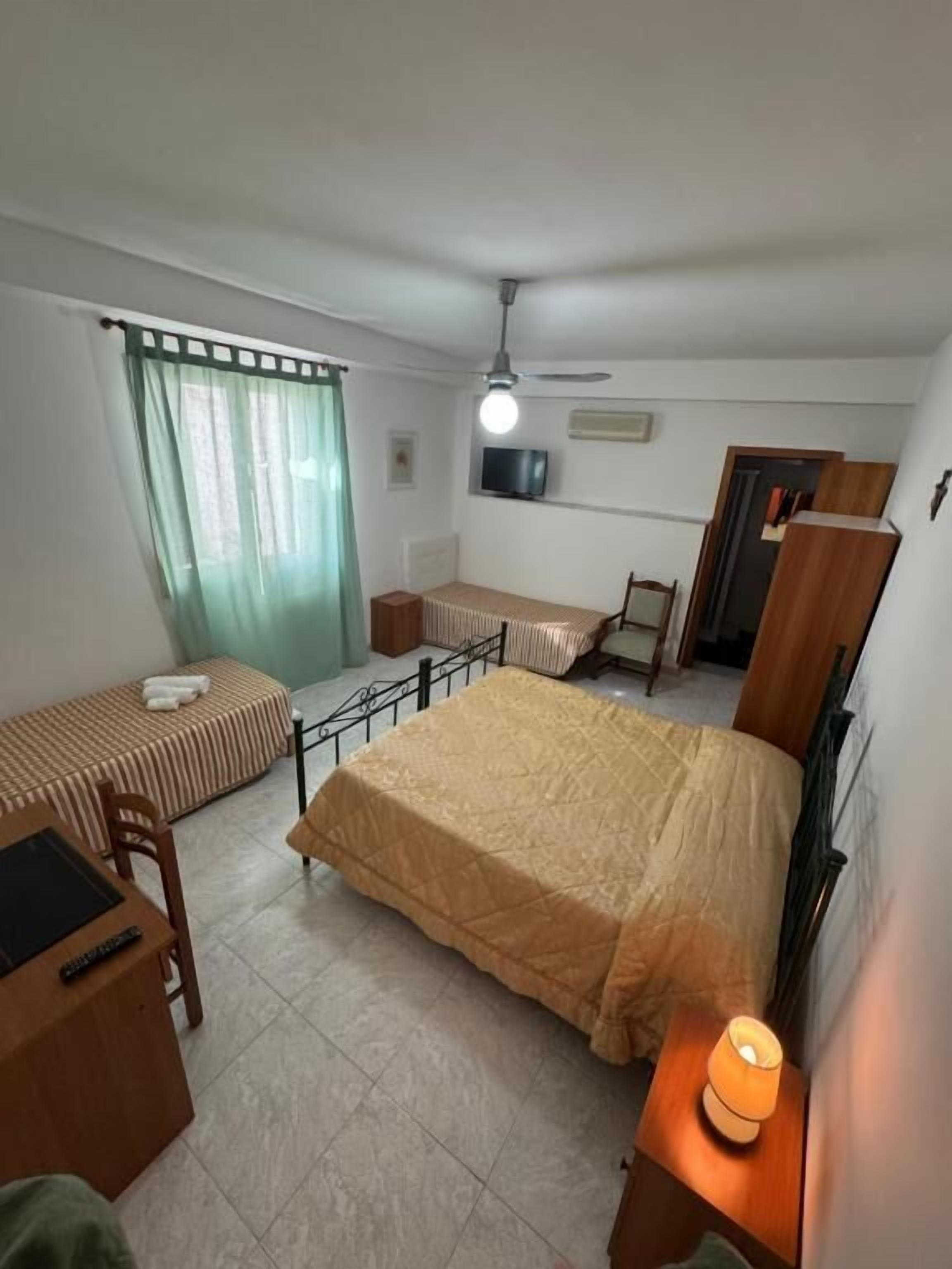A bedroom with a bed, a desk, a chair, a TV, a ceiling fan, and a window with curtains.