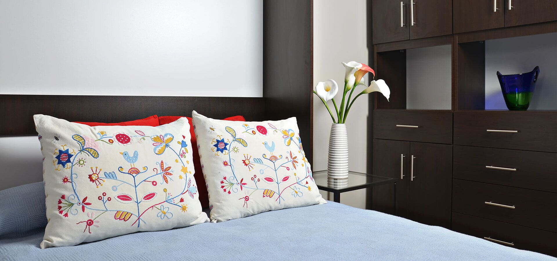 A bed with two decorative pillows, a vase with flowers, and a dark wooden dresser with drawers.