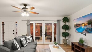 A living room with a gray sofa, a white coffee table, a ceiling fan, and a TV displaying a pool scene. - Sip & Swim | Heated Pool • Hot Tub • Games (Tucson)