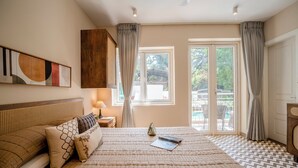 A bedroom with a large bed, a headboard, a bedside table with a lamp, a window with curtains, and a view of the outdoors. - Staymaster Solintra 2BHK C402 (Verem)