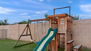 A wooden play structure with a slide and swings, set against a brick wall and grassy yard.