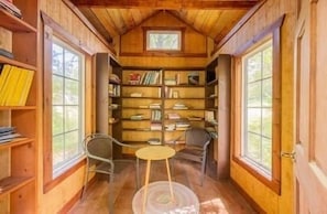 Ein Holzblockhaus-Innenraum mit Bücherregal, zwei Stühlen, einem kleinen Tisch und großen Fenstern.