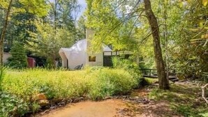 A natural setting with a small stream, dense greenery, and a modern building structure. - Creekside Dome with Firepit (Carroll County)