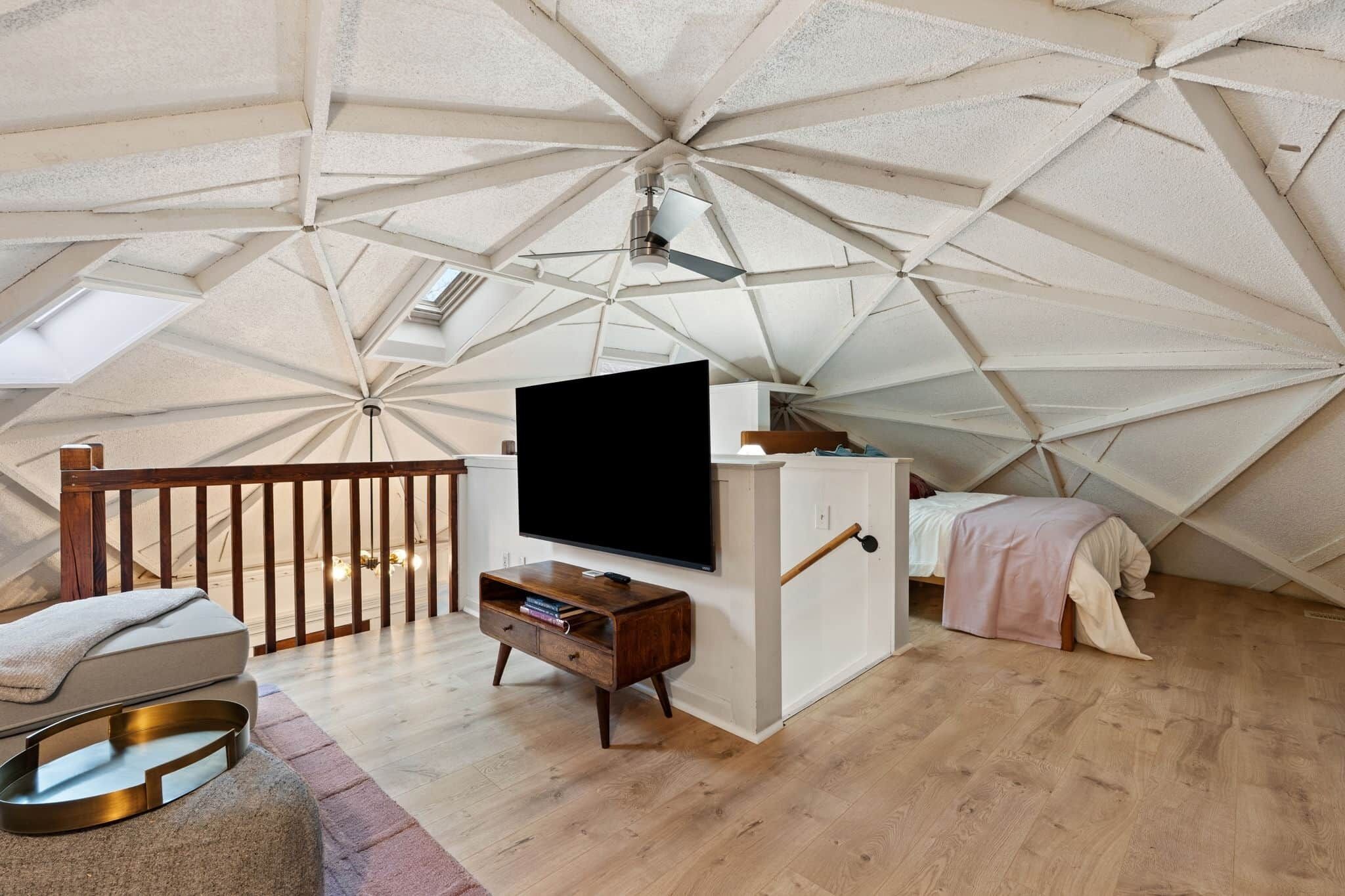A room with a sloped ceiling, a flat-screen TV mounted on the wall, a wooden bench, a bed with a pink blanket, and a small table with a tray.