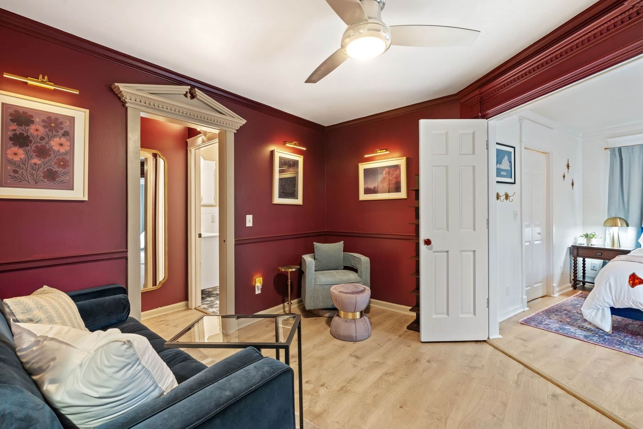 Una habitación con una pared de color rojo oscuro, un ventilador de techo, un sofá, una silla, una mesita y una puerta que da a otra habitación.