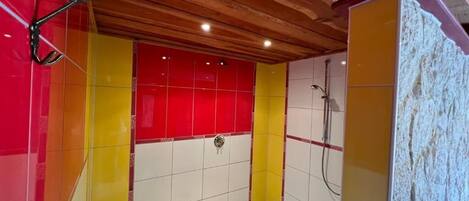 Un baño con techo de madera, paredes cubiertas de baldosas en rojo, amarillo y blanco, y una zona de ducha.