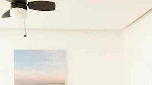 Een slaapkamer met een bed, kussens, een plafondventilator en een schilderij van een strand.