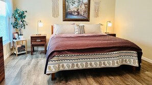 Quarto com cama grande, mesas de cabeceira, um criado-mudo, um vaso com planta e um quadro na parede.