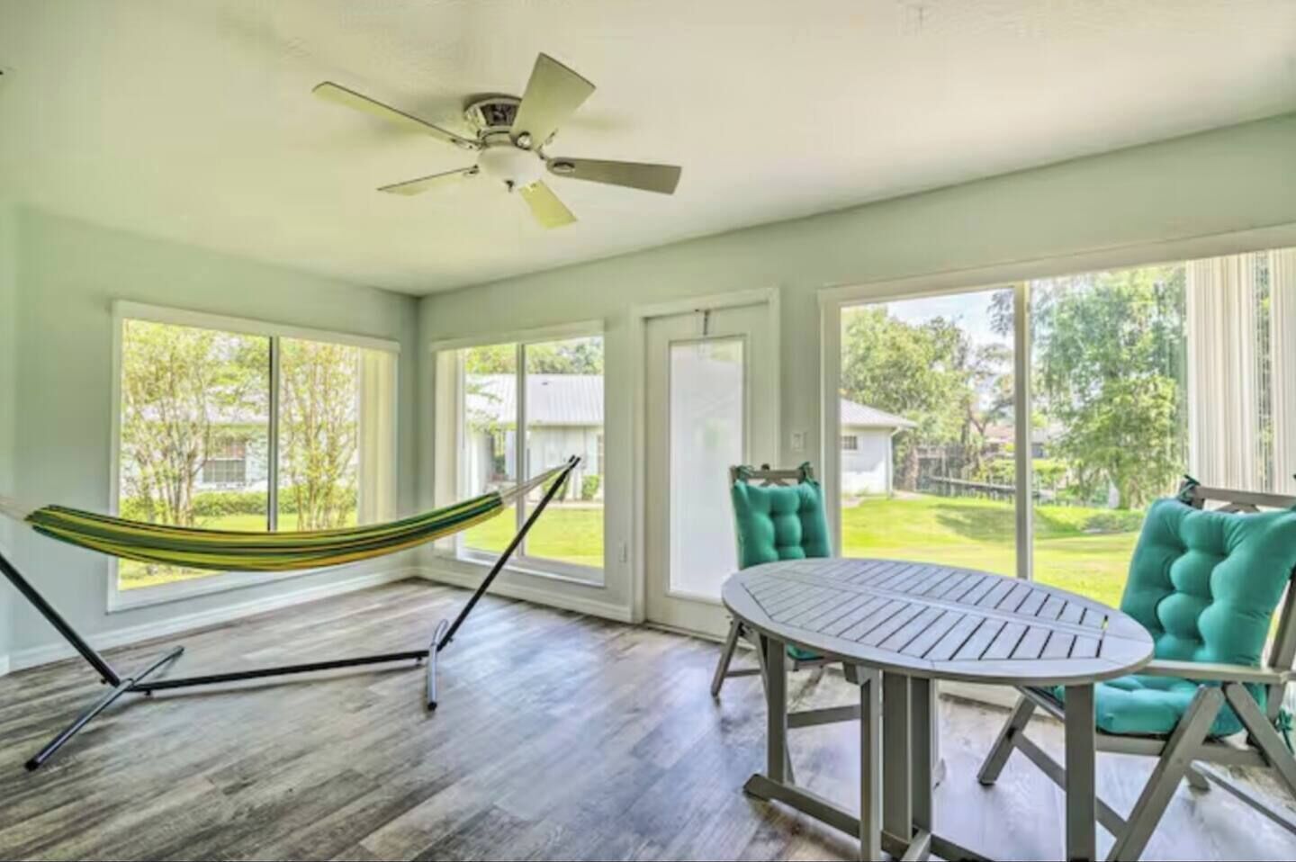 A room with a hammock, a round table, teal chairs, and large windows.