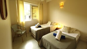Ein Zimmer mit zwei Betten, einem Sessel, einem Fenster mit Vorhängen und einer Wandleuchte.