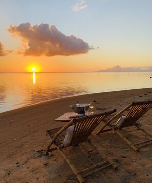 A beach scene with a sunset, a table with a pot and a glass, and two bamboo chairs.
