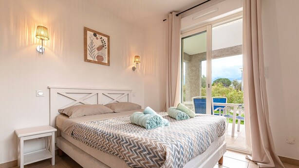 Ein Schlafzimmer mit Bett, Nachttisch, Wandlampe und Blick ins Freie.