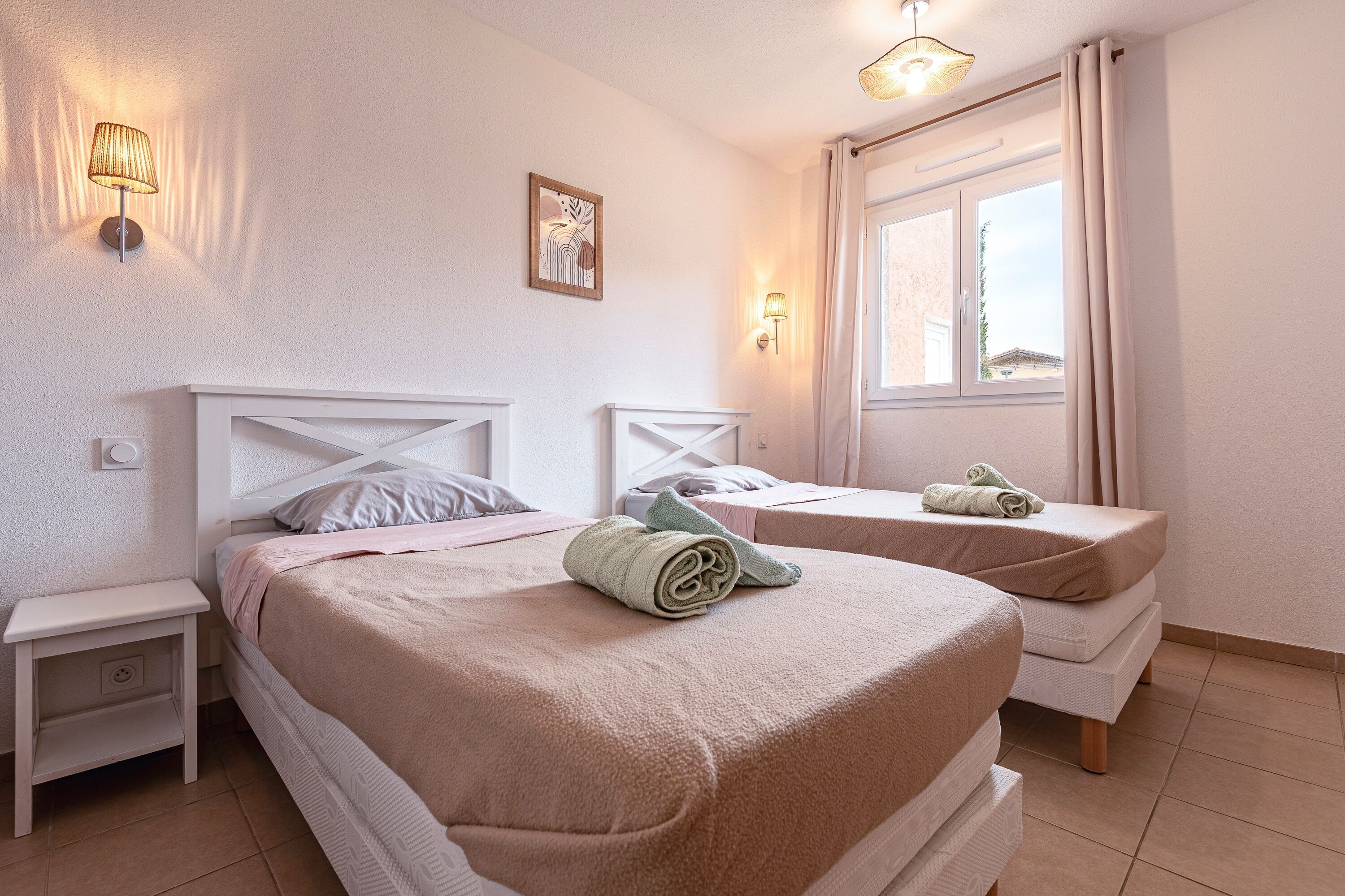 Una camera d'albergo con due letti, un comodino, una lampada a parete, una finestra con tende e un piccolo quadro appeso al muro.