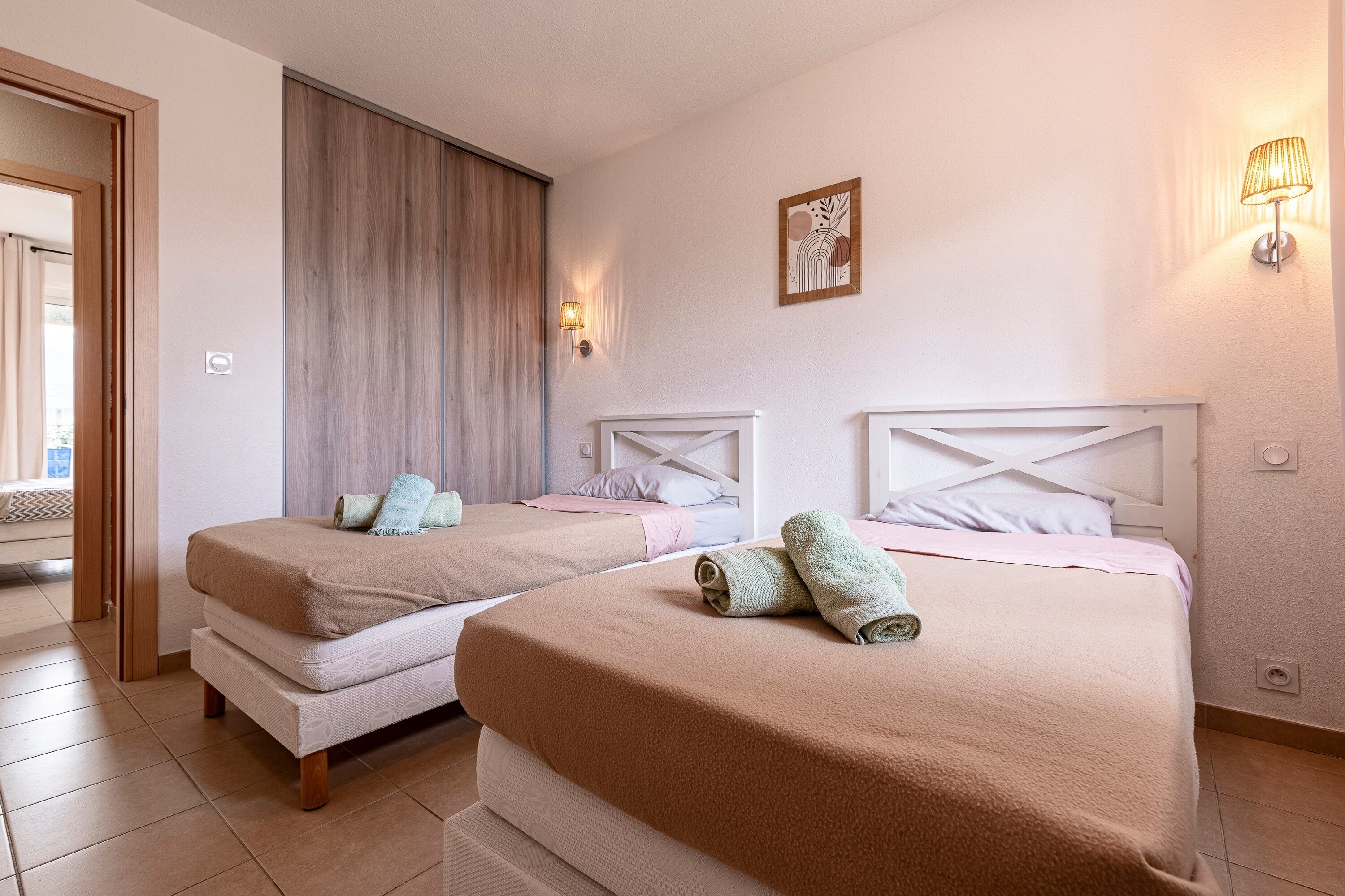 A hotel room with two beds, a wooden door, a wall-mounted light, and a framed picture.