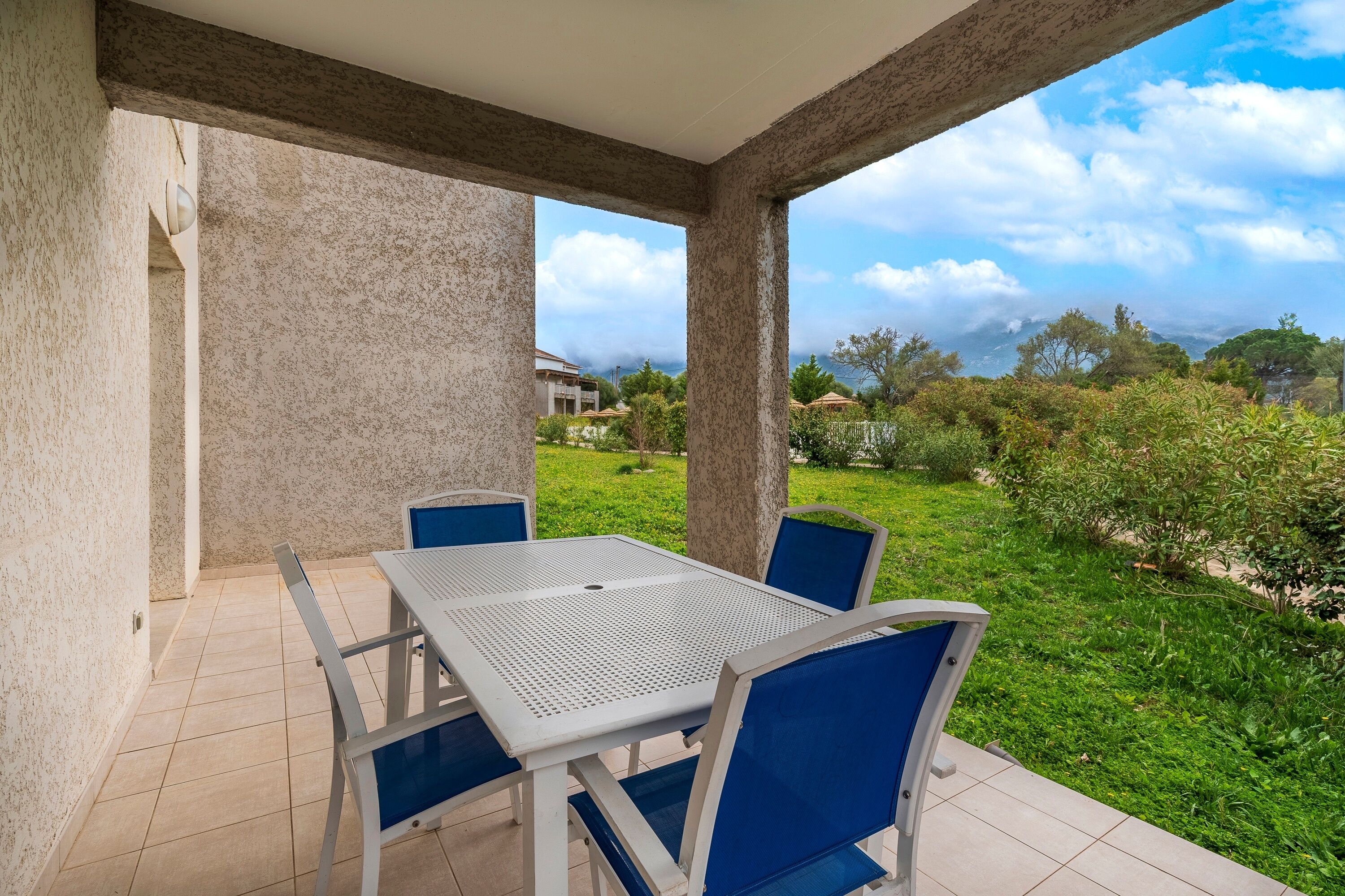 An outdoor patio with a white table and blue chairs, surrounded by a garden and a view of the landscape.