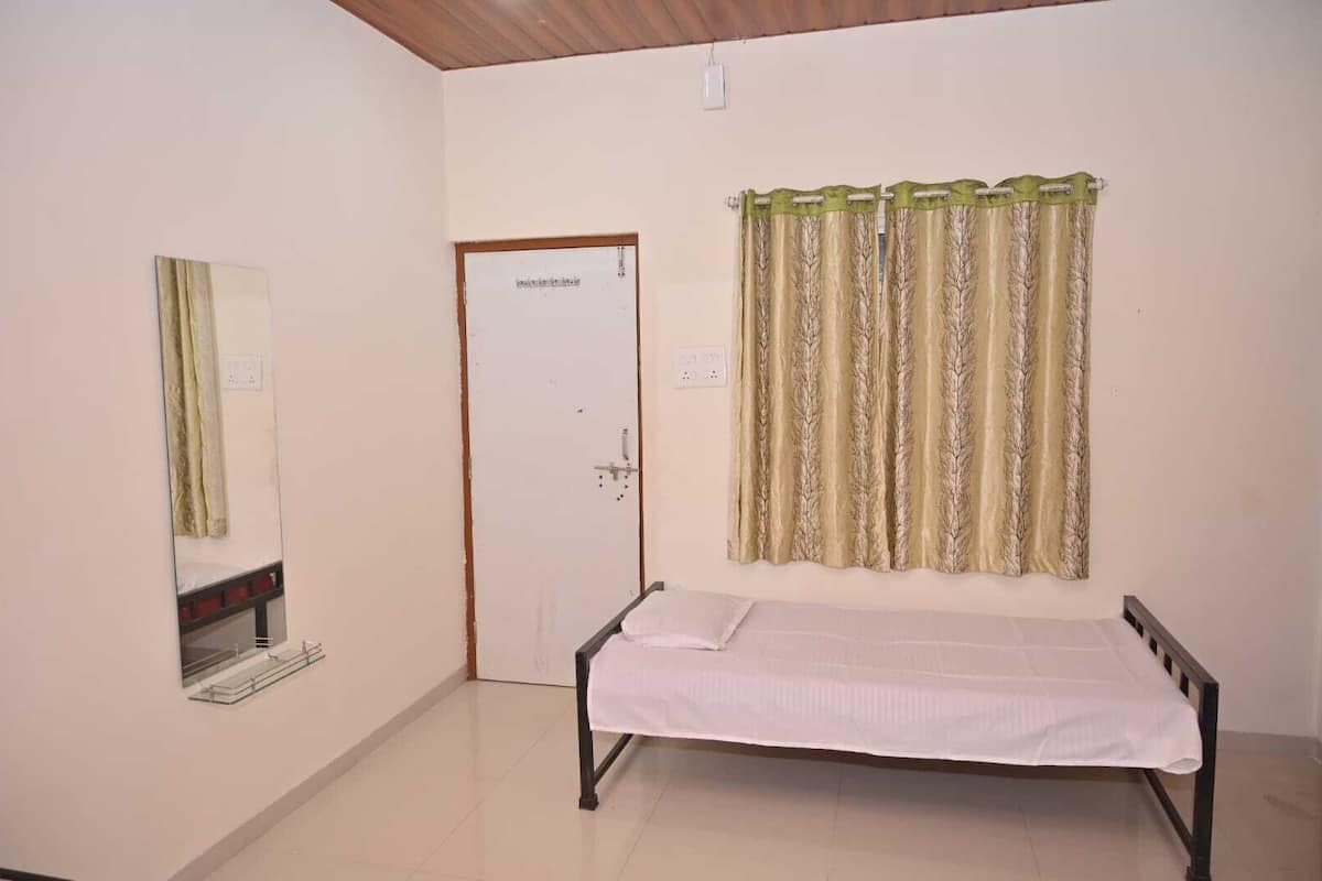 A single bed with a white mattress and a headboard, a mirror on the left wall, a door with a lock, and a window with patterned curtains.