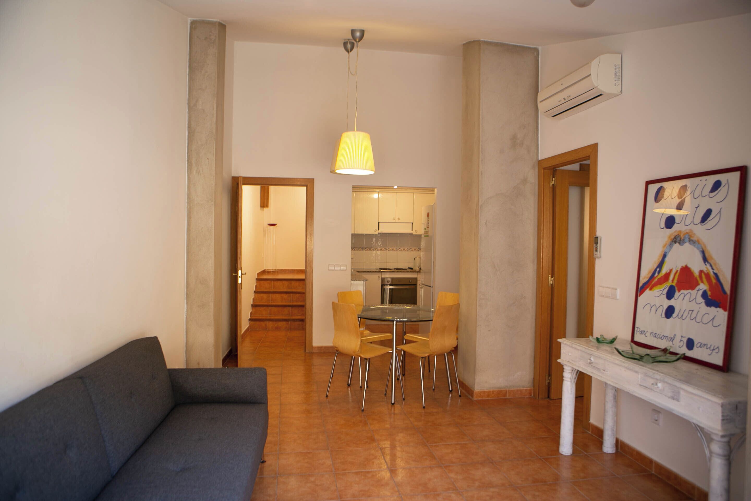 A living room with a sofa, a dining area with chairs, a table, a hanging light fixture, and a kitchen in the background.