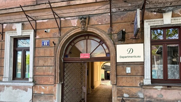 Ein Gebäudeeingang mit einem Metallgitter, einem Schild für 'Dapartmenty.eu' und einem kleinen Fenster mit Blick.