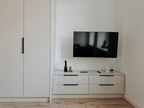 Habitació minimalista amb un televisor de pantalla plana al mur, un armari blanc amb calaixos i un terra de fusta.