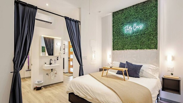 A modern bedroom with a bed, bedside tables, a mirror, a green wall with a neon sign, and a bathroom with a sink and mirror.