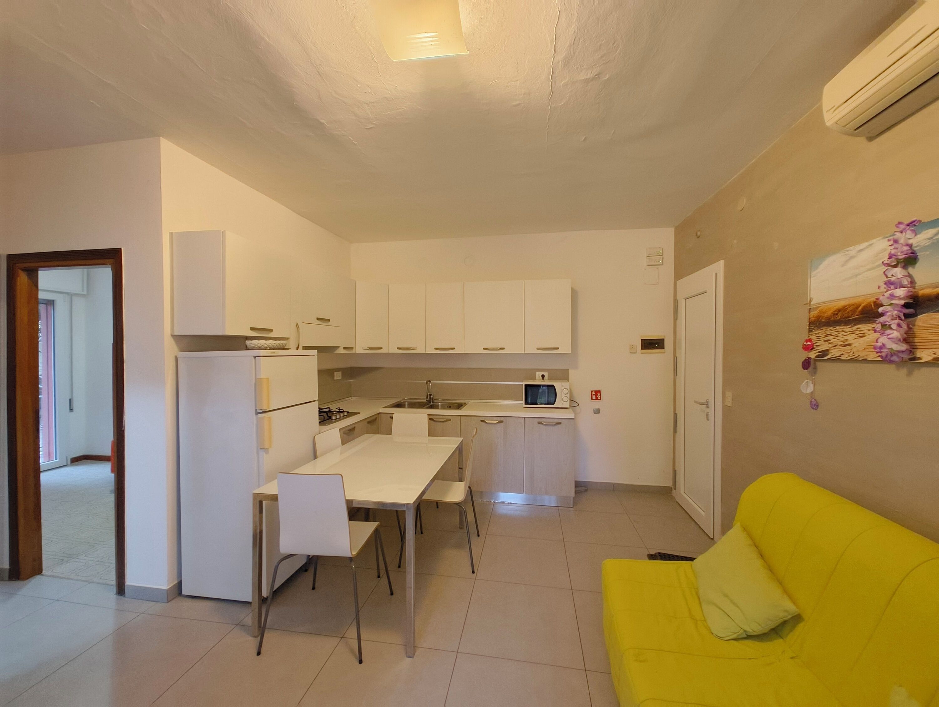 A compact kitchen with white cabinets, a dining table with chairs, a yellow sofa, and a small window.