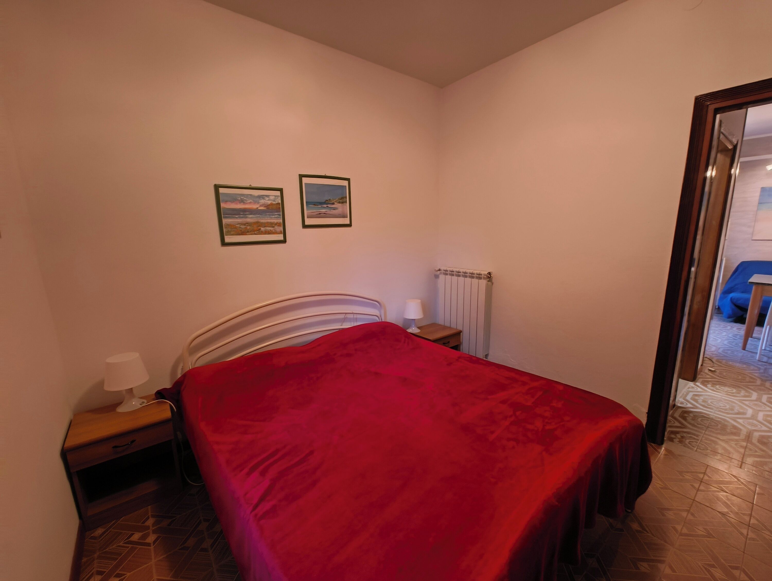 A bedroom with a bed, a nightstand, a lamp, a radiator, and two framed pictures on the wall.
