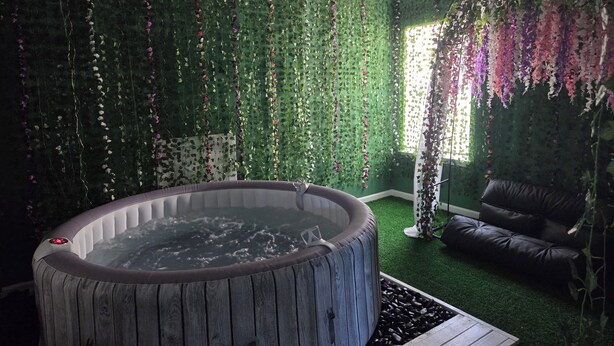 A room with a hot tub, a green wall with hanging flowers, a black leather sofa, and a wooden deck.