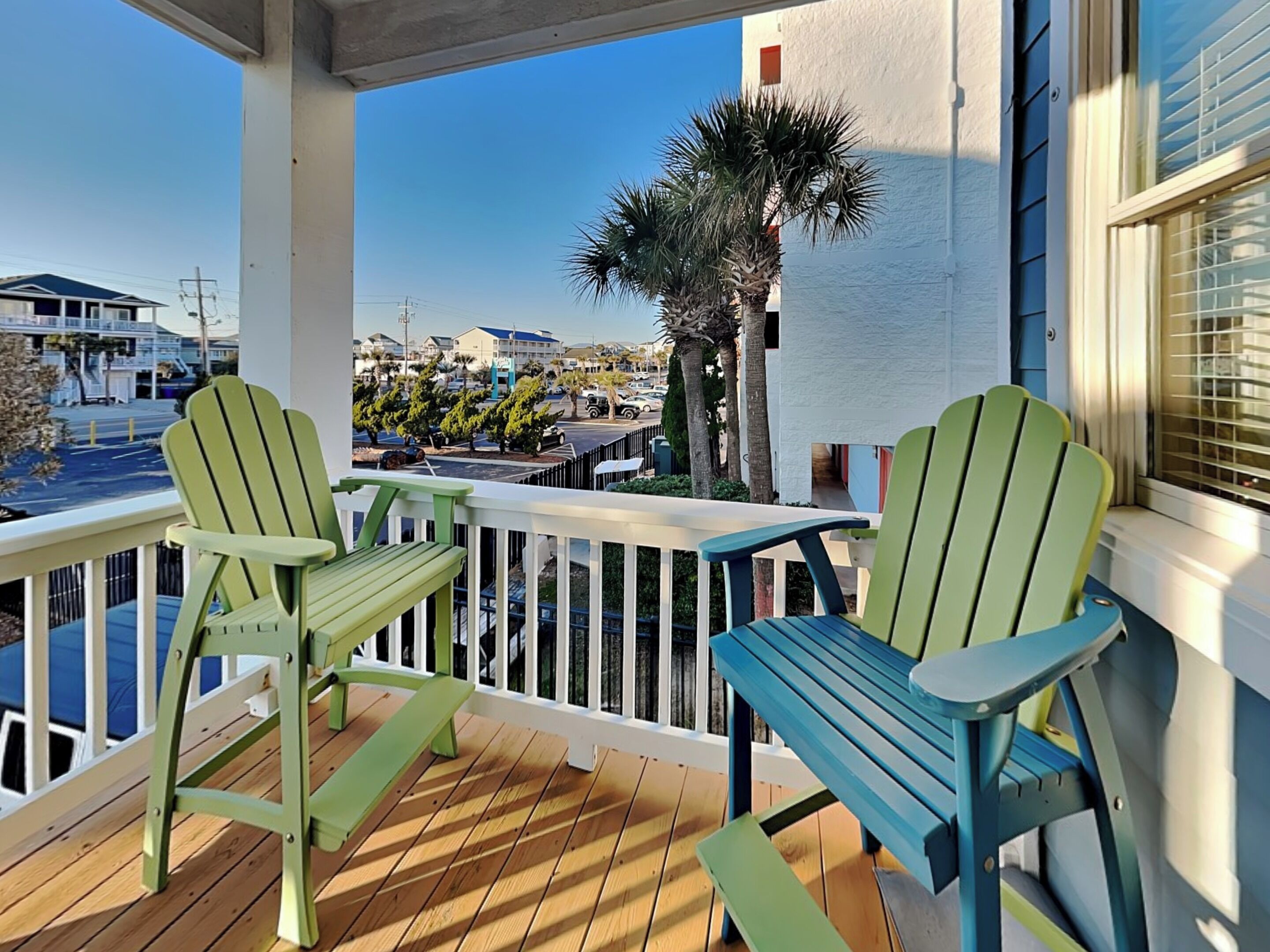 Un balcon avec deux fauteuils Adirondack, une terrasse en bois et une vue sur une rue bordée d’immeubles et de palmiers.