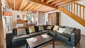 A cozy living room with a sectional sofa, wooden beams on the ceiling, a dining area, and a staircase.