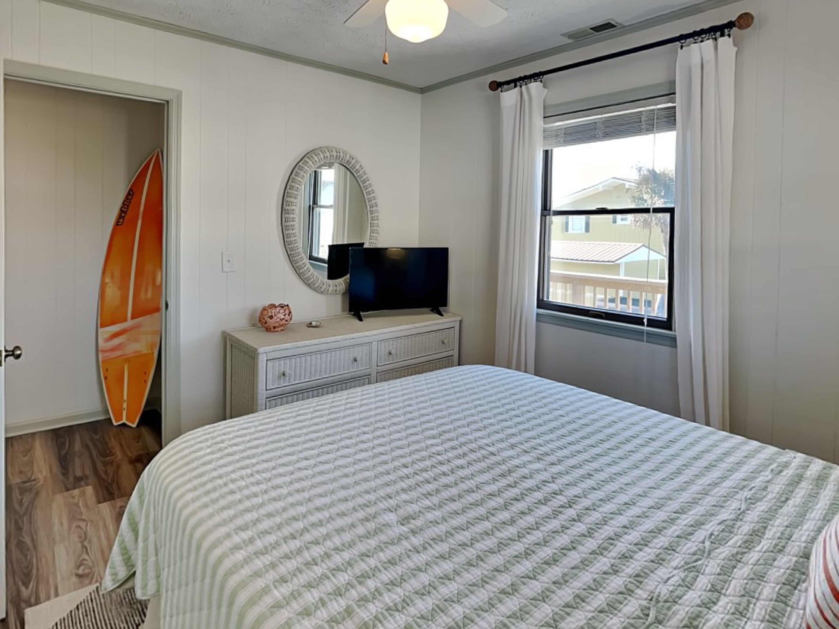 Een slaapkamer met een bed, een televisie op een dressoir, een surfplank en een raam met gordijnen.